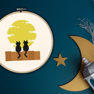 Puede incluir: Aro de bordado de punto de cruz con un sol amarillo y dos gatos negros sentados en una repisa marrón. El fondo es blanco.