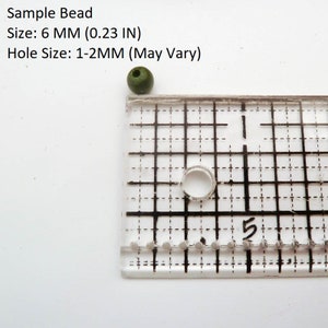 May include: A dark green round bead with a hole in the center, measured against a ruler. The bead is 6 mm (0.23 inches) in diameter. The hole size is 1-2 mm.  Sample Bead. Size: 6 MM (0.23 IN). Hole Size: 1-2MM (May Vary).