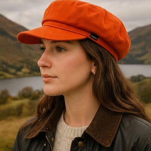 May include: Orange newsboy cap with a small brim. The cap has a structured shape and a small black label on the side. The cap is worn with a brown jacket and a cream-colored sweater.