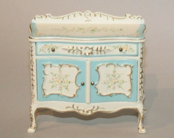 Puede incluir: Un armario de madera pintado de blanco y azul con detalles dorados y diseños florales. El armario tiene dos puertas y un cajón. El armario está sobre cuatro patas.