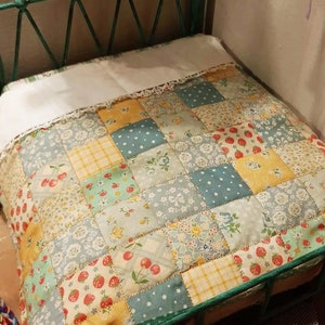 Op de afbeelding: Een groen metalen bedframe met een wit en blauw patchwork dekbed. Het dekbed heeft verschillende bloem- en geometrische patronen.