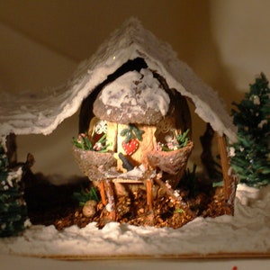 May include: A miniature winter scene with a small house made from a nut shell, covered in snow, with a snowy roof and a small tree in front. The house has a small door and windows, and a red heart on the front. The scene is set on a brown base with a white snowy surface.