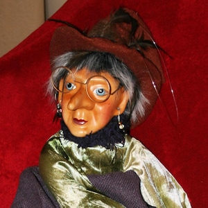 May include: A green and purple witch doll with a brown hat and glasses, riding a broomstick with a brown bag hanging from it.