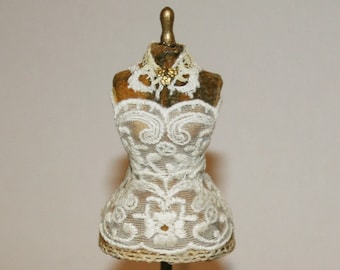 May include: A miniature gold-toned dress form with a white lace bodice and a gold-toned stand with three legs.