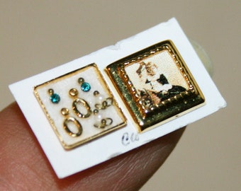 May include: A white card with two gold-framed earrings. One earring has a design of small white and blue circles. The other earring has a picture of a woman in a white dress.