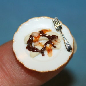 Puede incluir: Plato de postre en miniatura con borde festoneado y ribete dorado. El plato contiene una pequeña porción de panqueques rociados con salsa de chocolate y caramelo. Un tenedor plateado diminuto descansa en el plato, sugiriendo un escenario de casa de muñecas.