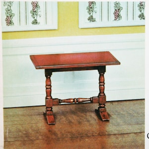 May include: A miniature wooden trestle table with a dark brown finish. The table has a rectangular top and four turned legs. The table is sitting on a wooden floor. The text "TRESTLE STAND/Connecticut, Circa 1770-90" is printed on the bottom of the image.