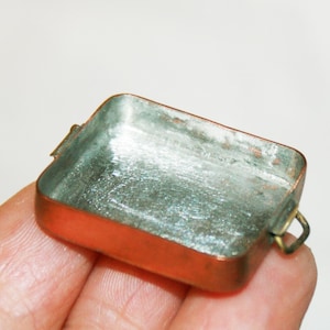 Puede incluir: Un pequeño recipiente rectangular de color cobre con un interior deslustrado de color plateado. El recipiente tiene un pequeño asa curvada en un lado. El objeto se sostiene en la mano de una persona.