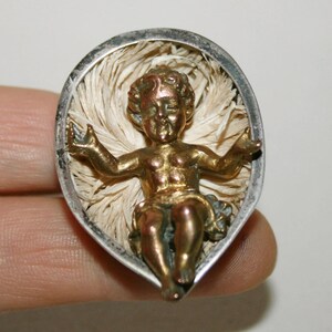 Pode incluir: Um pingente prateado e dourado com uma figura de bebê Jesus em um presépio. O presépio é feito de fibras brancas e é cercado por uma moldura prateada.