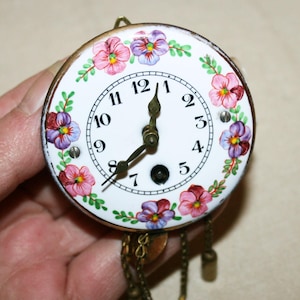 May include: A round clock face with a white background, black Arabic numerals, and dark metal hands. The border is decorated with hand-painted pink and purple pansy flowers and green leaves. Two metal chains with small weights hang from the bottom.