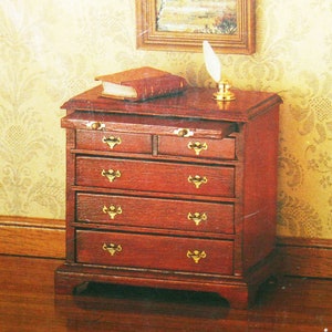 May include: A wooden chest of drawers with five drawers and brass handles. The chest is a dark reddish brown colour and has a slightly distressed finish. A book and a quill pen are on top of the chest.