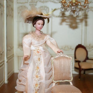 May include: A miniature doll in a cream-coloured Victorian-style gown and hat, holding a small chair. The doll is in a detailed room setting with a chandelier, two chairs, and a patterned rug. The room has ornate wall panelling.
