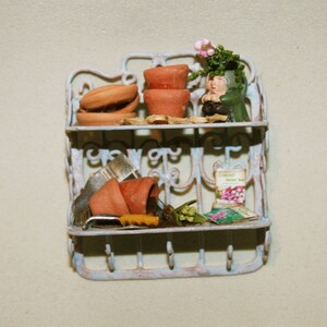 May include: A white metal shelf with two tiers, decorated with floral designs. The top tier has three terracotta flower pots, one with a small green plant and a miniature figurine. The bottom tier has a terracotta pot, a small gardening tool, and a packet of flower seeds. The shelf is against a light gray wall.