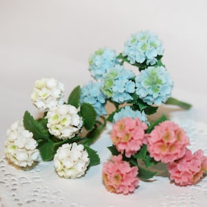 Op de afbeelding: Drie takjes kunstbloemen met witte, lichtblauwe en roze bloemen. De bloemen zijn gerangschikt op een wit kantonderzetter.