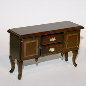 May include: A small, dark brown wooden cabinet. It features two drawers with gold-coloured handles and a single door. The cabinet has curved legs and a darker brown top. The dimensions are approximately 15 cm in length, a miniature piece of furniture.