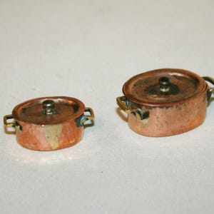 May include: Two copper pots with brass handles and lids. The pots are oval shaped and have a shiny finish.