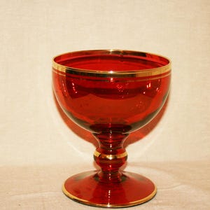 May include: A red glass goblet with gold trim. The goblet has a rounded bowl, a decorative stem, and a circular base. The gold rim and base add a touch of elegance to the vintage glassware. The goblet is suitable for serving wine or other beverages.