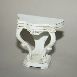 May include: A small, white, ornate table with a curved top and decorative legs. The table has a classic design with floral accents and a distressed finish, suggesting an antique or vintage style. The table is approximately 12 inches tall.