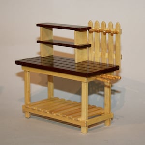 May include: A miniature wooden potting bench featuring a dark brown work surface, a light natural wood frame, and a slatted lower shelf. The unit includes a two-tier upper shelf, a small picket fence back panel, and several small wooden pegs.