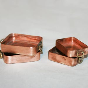 Puede incluir: Cuatro pequeños recipientes rectangulares de cobre con asas color latón. Dos recipientes están apilados y dos están uno al lado del otro. El cobre tiene un tono marrón rojizo.