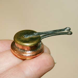 Może przedstawiać: Miniaturowa miedziana patelnia z mosiężną pokrywką i dwoma małymi metalowymi uchwytami. Patelnia ma około 5 cm średnicy i leży na palcu.
