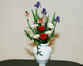 May include: A white ceramic vase with a classic design holds a bouquet of colorful flowers. The bouquet includes red roses, white daisies, pink lilies, and blue irises.