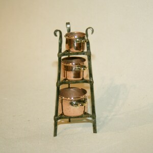 May include: A set of four copper pots with handles, stacked on a black metal stand with decorative scrolls. The pots are nested inside each other, with the largest pot at the bottom and the smallest pot at the top.