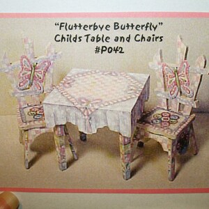 Op de afbeelding: Een miniatuur houten tafel en twee stoelen, geschilderd in pastelkleuren met vlindermotieven. De tafel heeft een roze-wit geblokt tafelkleed. De tekst luidt "Flutterbye Butterfly" Childs Table and Chairs #P042.