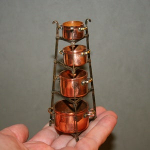 May include: A set of four copper pots stacked on a metal stand. The pots are of varying sizes and have handles. The stand is made of black metal and has a decorative design.
