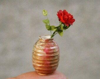 May include: A small, gold-colored ceramic vase with a textured surface, holding a single red flower with green leaves.