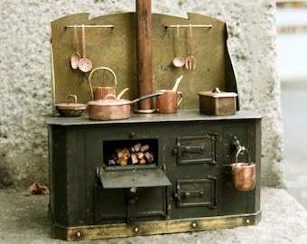 May include: A miniature cast iron stove with a copper chimney and a brass backsplash. The stove has an open oven door revealing a small pile of wood logs. There are several copper pots and pans hanging from hooks on the backsplash.