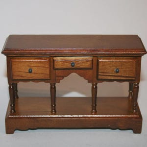 May include: A miniature wooden desk with three drawers and a lower shelf. The desk is stained a rich brown colour and features small black knobs on the drawers. The legs are turned and the overall design is ornate.