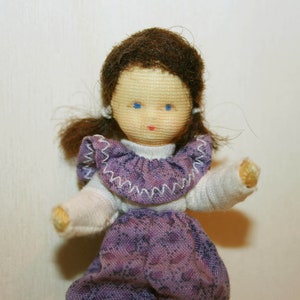 May include: A handmade doll with brown hair, blue eyes, and a purple and white dress. The doll has a simple, sweet expression.