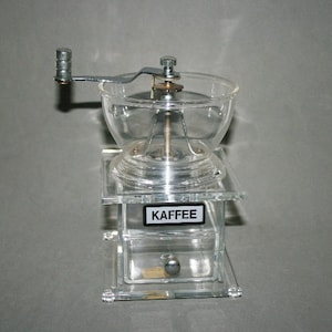 Puede incluir: Un molinillo de café de plástico transparente con manivela y cajón. El molinillo tiene una etiqueta que dice "Kaffee".