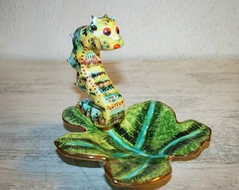 Puede incluir: Una figurita de caballito de mar de cerámica con rayas amarillas, azules y marrones, sentada sobre un plato verde en forma de hoja con detalles dorados.