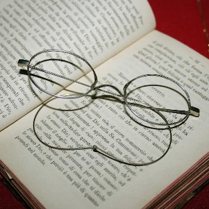 May include: An open book with text and a pair of round, wire-rimmed eyeglasses resting on the pages. The glasses are silver-toned, and the book has aged, yellowed pages.