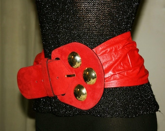 May include: A red belt with a large, suede-like buckle featuring three gold-colored circular accents. The belt's main strap is a vibrant red, and it is wrapped around a black garment. The buckle is a prominent design element.