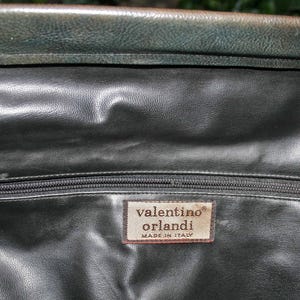 May include: Close-up of a dark green leather bag interior with a zipper closure. A rectangular tag reads "valentino orlandi MADE IN ITALY". The leather has a textured appearance.