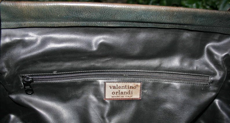 May include: Close-up of a dark green leather bag interior with a zipper closure. A rectangular tag reads "valentino orlandi MADE IN ITALY". The leather has a textured appearance.