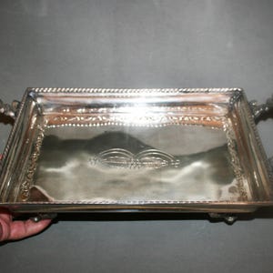 May include: A rectangular silver-toned metal serving tray with ornate, curved handles on each side. The tray features a raised, rope-style border and a decorative engraved pattern in the center of the flat base, supported by small feet.