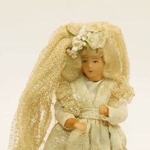 May include: A white fabric doll wearing a white lace dress and veil with a floral crown. The doll has a painted face with blonde hair.