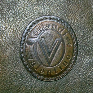 May include: Close-up of a dark green leather surface with a circular, embossed emblem. The emblem features the name "Valentino" and a stylized "V" within a circular frame, stitched to the leather.