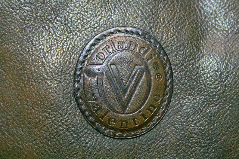 May include: Close-up of a dark green leather surface with a circular, embossed emblem. The emblem features the name "Valentino" and a stylized "V" within a circular frame, stitched to the leather.