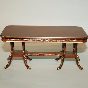 Puede incluir: Una mesa marrón en miniatura con una tapa rectangular y adornos dorados. La mesa tiene dos estantes y patas decorativas con detalles dorados. La mesa mide aproximadamente 30 cm de largo.