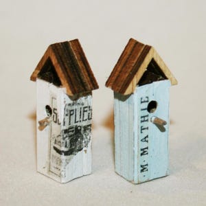 May include: Two miniature birdhouses with wooden roofs and light-colored bodies. One is painted white with a partially visible label, the other is light blue with the text "M. MATHIE". Each has a small wooden perch.