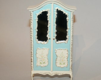 Puede incluir: Un pequeño armario ornamentado en azul claro y blanco. Cuenta con dos puertas con espejos, detalles florales y adornos dorados. El armario tiene una parte superior curvada y se apoya en cuatro delicadas patas. Altura aproximada: 60 cm.