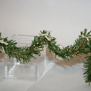 May include: A green garland with gold and white accents. The garland is decorated with small gold balls and white bows. The garland is draped over a clear glass cube.