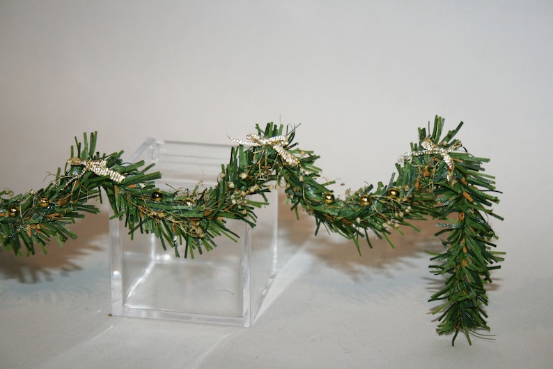 May include: A green garland with gold and white accents. The garland is decorated with small gold balls and white bows. The garland is draped over a clear glass cube.