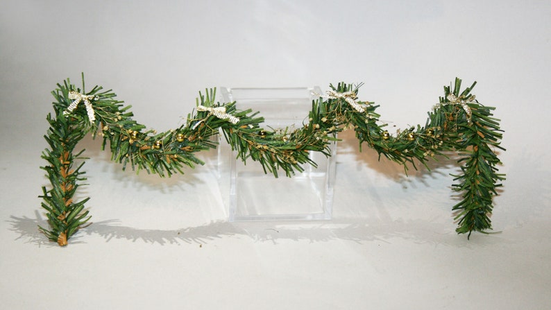 May include: A green garland with gold bells and white bows. The garland is draped over a clear acrylic block.