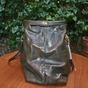 May include: A dark green leather handbag with a shoulder strap and gold-colored hardware. The bag has a soft, slouchy design and a top handle. The background features green foliage and a wooden surface.